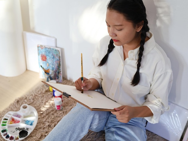 Jovem feliz sentada em casa e desenhando com uma paleta de tinta e um artista de pintura com pincel