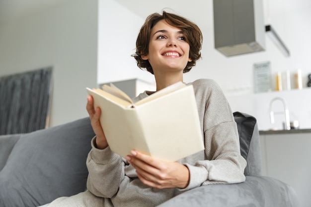 Jovem feliz sentada em um sofá em casa, lendo um livro