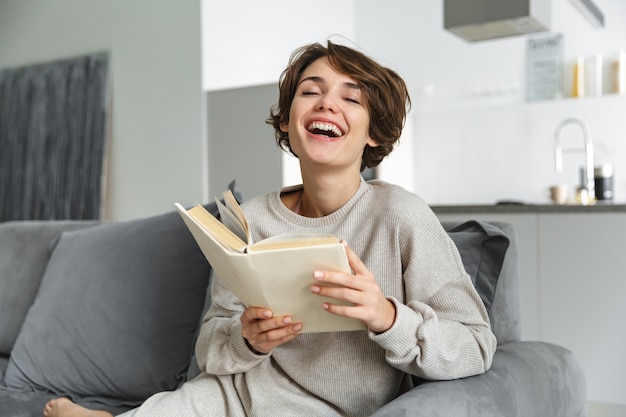 Jovem feliz sentada em um sofá em casa, lendo um livro