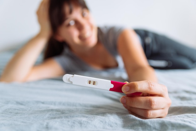 Jovem feliz sentada na cama olhando para um teste de gravidez positivo fundo desfocado