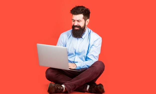 Jovem feliz sentado no chão com e usando o computador laptop sobre fundo vermelho.