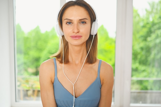 Jovem feliz sorridente linda slim fit mulher azul sportswear ouvindo música em grandes fones de ouvido brancos em casa