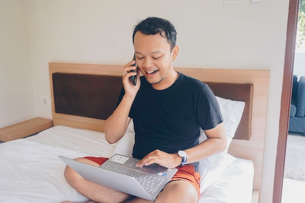 Jovem feliz trabalhando na cama segure o laptop de joelhos e falando ao telefone