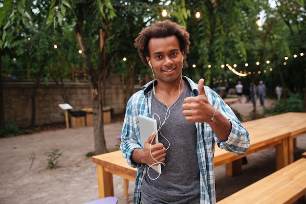 Jovem feliz usando fones de ouvido segurando um tablet e mostrando o polegar para cima