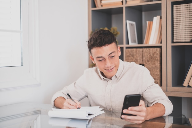 Jovem feliz usando seu smartphone em casa navegando na net, fazendo lição de casa, se divertindo sozinho dentro de casa. Adolescente do sexo masculino com telefone aproveite o tempo livre, estudando e trabalhando.