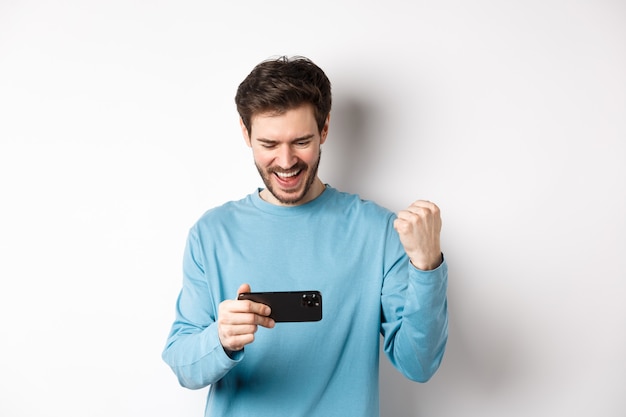 Jovem feliz vencendo no videogame no smartphone, olhando para a tela do celular e dizer sim, fazendo o punho bater em comemoração, atingir a meta online, em pé sobre um fundo branco