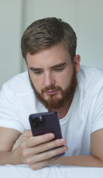 Jovem feliz veste camiseta branca segura usa celular navegando na internet deitado em repouso na cama