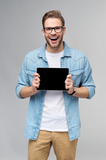 Jovem feliz vestindo uma camisa jeans em pé e segurando o teclado do tablet pc sobre a parede cinza do estúdio