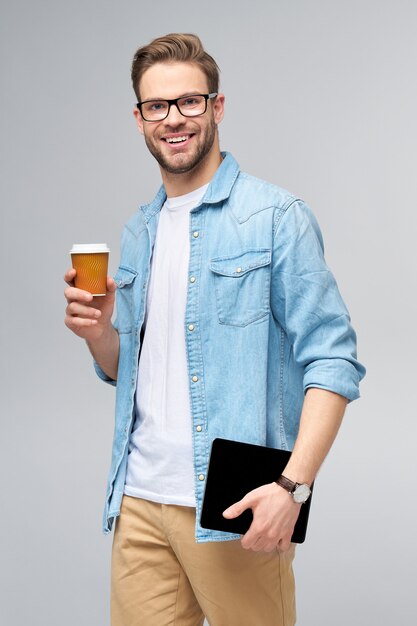 Jovem feliz vestindo uma camisa jeans em pé, usando o teclado do tablet pc e segurando uma xícara de café para ir