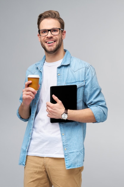 Jovem feliz vestindo uma camisa jeans em pé, usando o teclado do tablet pc e segurando uma xícara de café para ir