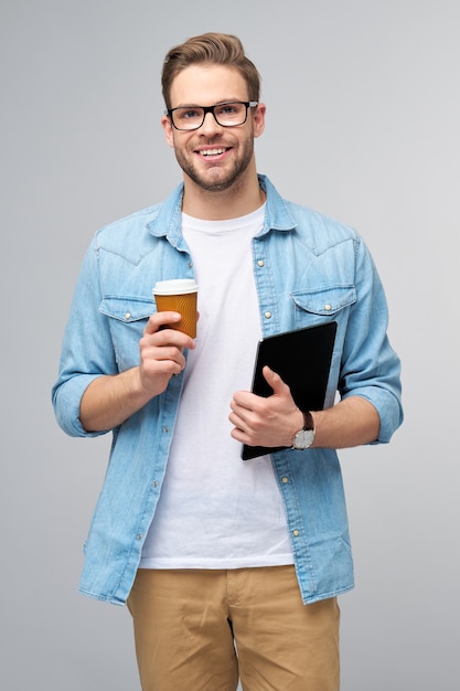 Jovem feliz vestindo uma camisa jeans em pé, usando o teclado do tablet pc e segurando uma xícara de café para ir