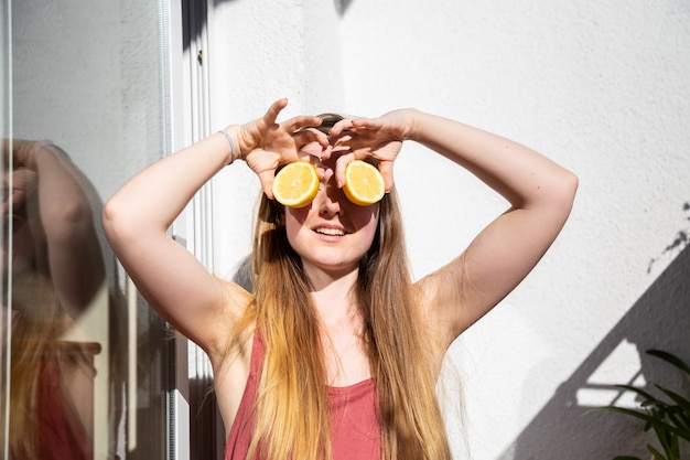 Jovem fêmea bonita feliz no vestido casual de verão, sentado no terraço e cobrindo os olhos com limões maduros