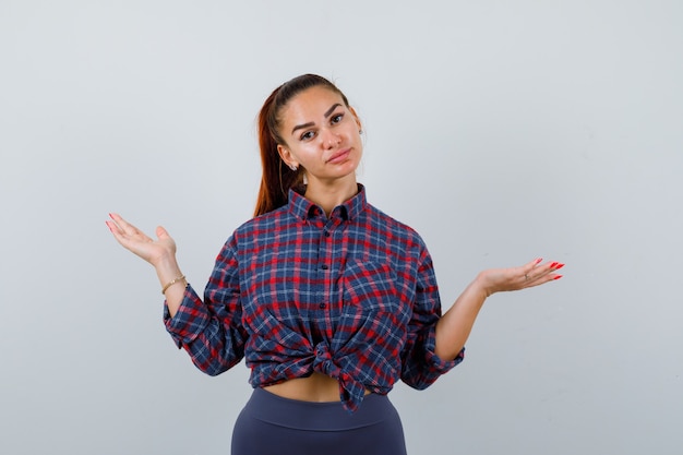 Jovem fêmea com camisa quadriculada, calça mostrando um gesto desamparado e parecendo sem noção, vista frontal.