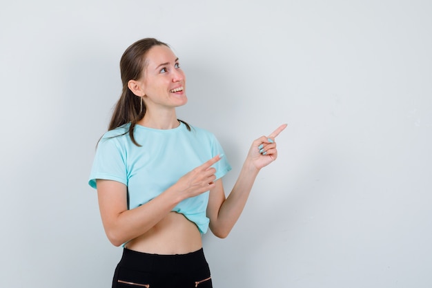 Foto jovem fêmea linda em t-shirt, apontando para o canto superior direito, olhando para cima e parecendo feliz, vista frontal.