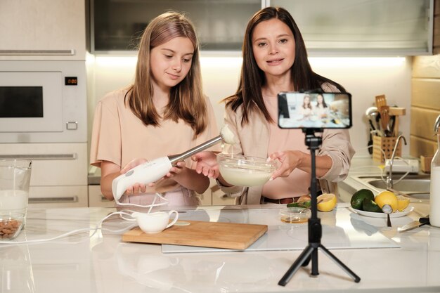 Jovem fêmea olhando para a câmera do smartphone e preparando sorvete caseiro na cozinha enquanto compartilha sua receita com o público online