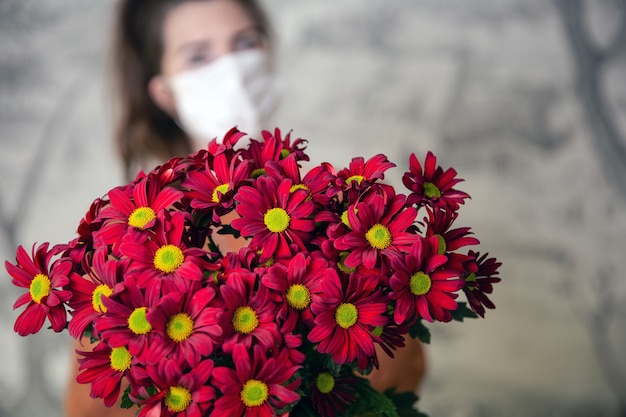 Jovem fêmea segurando um buquê de flores vermelhas margarida usando uma máscara protetora médica para coronavírus, dia dos namorados ou dia das mães, aniversário e conceito de Covid-19. design romântico