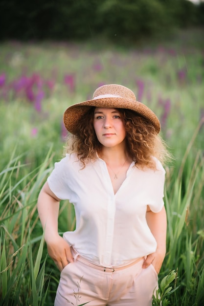 Jovem fica de camisa branca no campo de tremoços roxos e rosa Bela jovem com cabelo encaracolado e chapéu ao ar livre em uma flor de tremoços de prado Pôr do sol ou nascer do sol luz brilhante da noite