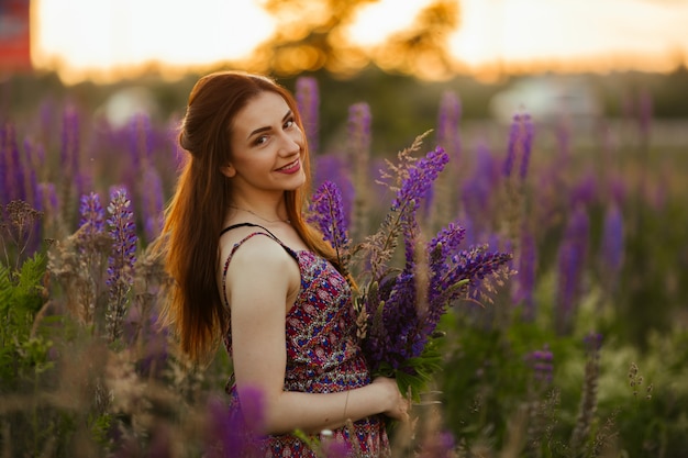 Jovem ficar no campo com vista para o campo de lavanda. Sorrindo despreocupada garota caucasiana em vestido, apreciando o pôr do sol