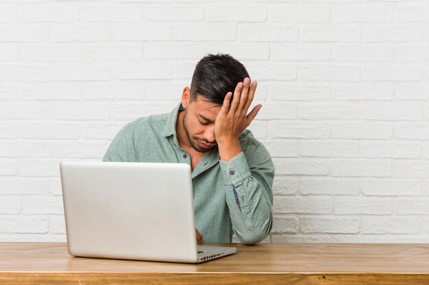 Jovem filipino sentado trabalhando com seu laptop, esquecendo algo, batendo na testa com a palma da mão e fechando os olhos.