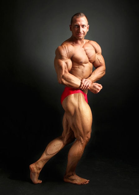 Jovem fisiculturista masculino posando, mostrando seus músculos do peito frontal, pernas e bíceps enorme, sobre fundo preto.
