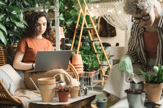 Jovem florista sentada no sofá digitando no laptop ela trabalhando no jardim junto com sua colega que está plantando plantas