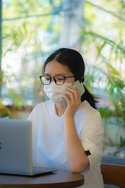 Jovem focada usando uma máscara médica usando laptop digitando no teclado sentado na cafeteria