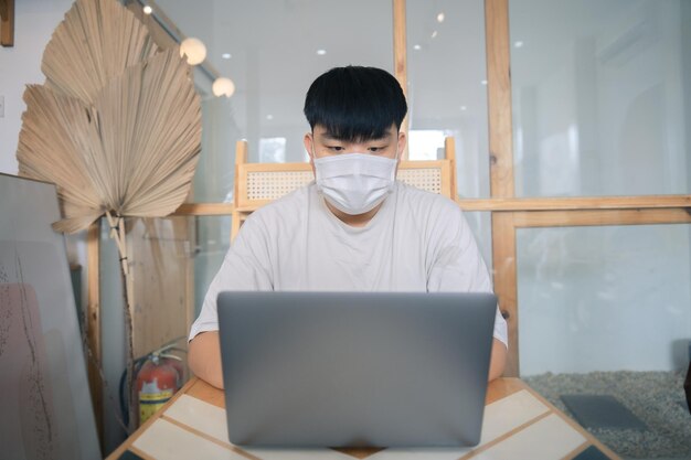 Jovem focado usando uma máscara médica usando laptop digitando no teclado escrevendo e-mail