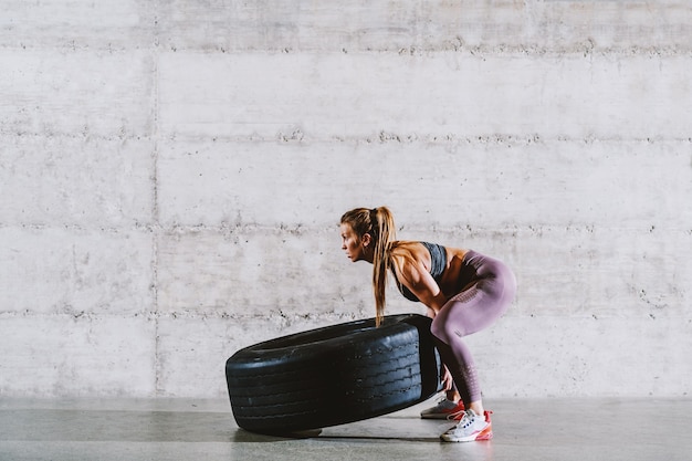 Jovem forte desportista caucasiana muscular em sportswear com rabo de cavalo lançando o pneu. conceito de estilo de vida saudável.