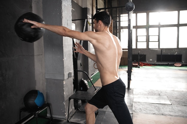 Jovem, forte, suado, focado, apto, homem musculoso, jogando medicine ball para cima na parede para treino de crossfit treino pesado no ginásio