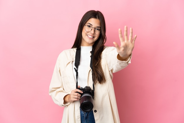 Jovem fotógrafa brasileira isolada em parede rosa contando cinco com os dedos