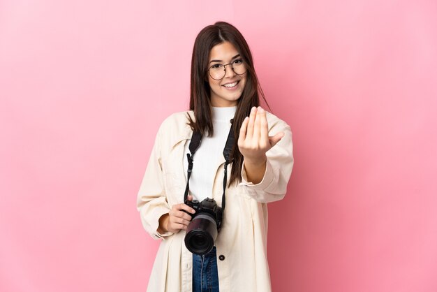 Jovem fotógrafa brasileira isolada na parede rosa convidando para vir com a mão. Feliz que você veio