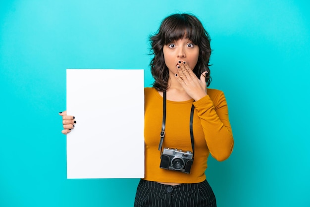 Jovem fotógrafa latina isolada em fundo azul segurando um cartaz vazio com expressão de surpresa