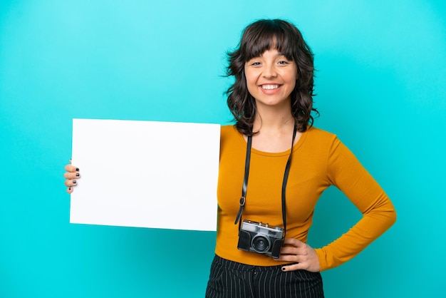 Jovem fotógrafa latina isolada em fundo azul segurando um cartaz vazio com expressão feliz