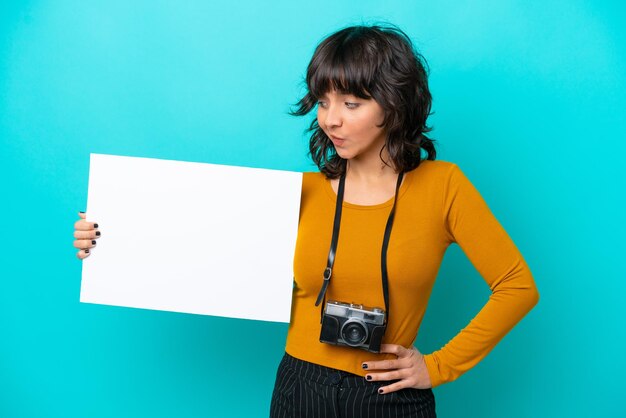 Jovem fotógrafa latina isolada em fundo azul segurando um cartaz vazio e olhando para ele