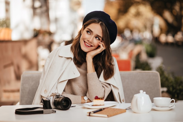 Jovem fotógrafo bonito, com um penteado ondulado escuro, boina vintage e sobretudo bege, descansando no terraço do café da cidade com chá, cheesecake, câmera e caderno na mesa