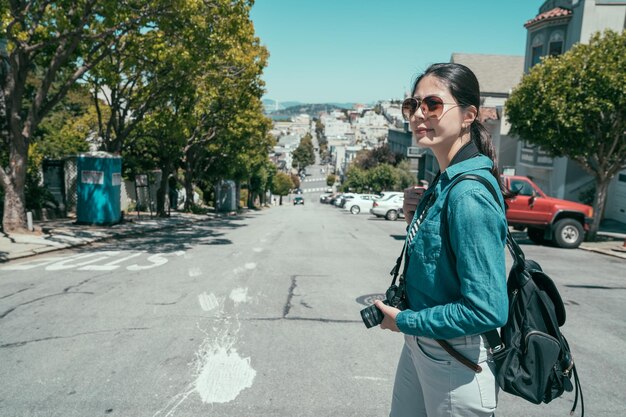 Jovem fotógrafo feminino tirando fotografia na câmera vintage ao ar livre na rua lombard. garota turista com mochila aproveitando a viagem de verão em são francisco, passeios turísticos na colina russa em clarão ensolarado