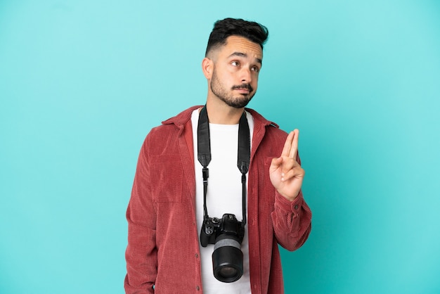 Jovem fotógrafo homem caucasiano isolado em um fundo azul com os dedos se cruzando e desejando o melhor