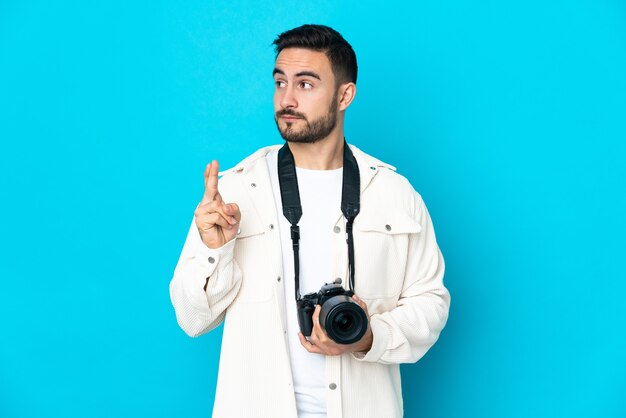 Jovem fotógrafo isolado em um fundo azul com os dedos se cruzando e desejando o melhor