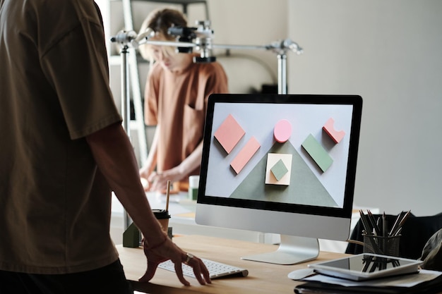 Jovem fotógrafo masculino de pé ao lado da mesa com monitor de computador