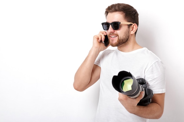 Jovem fotógrafo profissional de camisa branca segurando uma câmera digital pesada com uma lente longa e falando ao telefone com um cliente