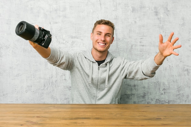 Jovem fotógrafo segurando uma câmera em uma mesa se sente confiante, dando um abraço para a câmera.