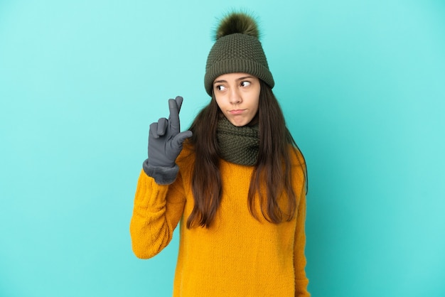 Foto jovem francesa isolada em um fundo azul com chapéu de inverno cruzando os dedos e desejando o melhor