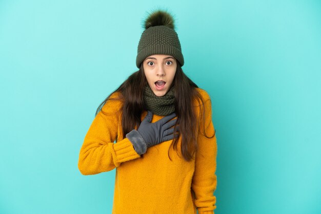 Jovem francesa isolada em um fundo azul com chapéu de inverno surpresa e chocada ao olhar para a direita