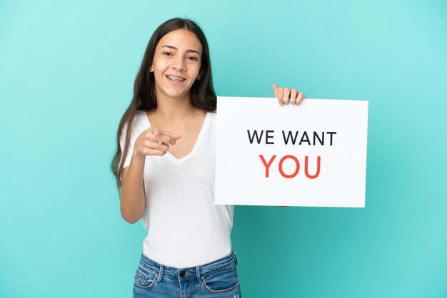 Jovem francesa isolada em um fundo azul segurando a placa We Want You e apontando para a frente