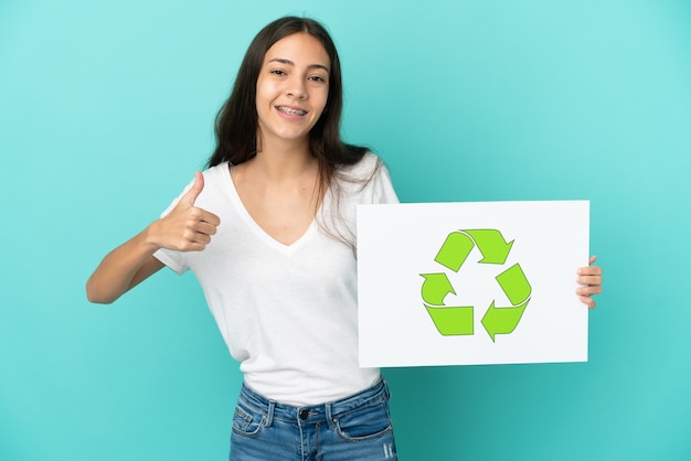 Jovem francesa isolada em um fundo azul segurando um cartaz com o ícone de reciclagem com o polegar para cima