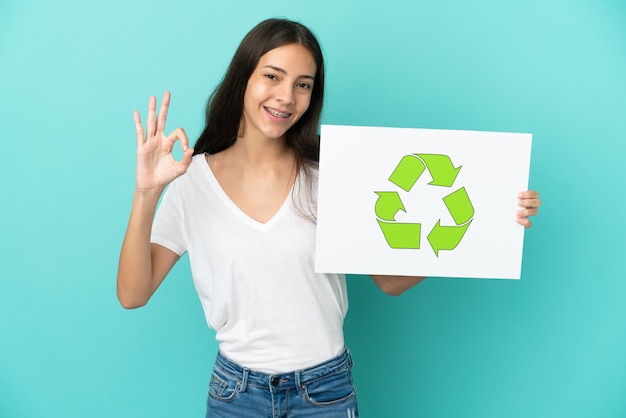 Jovem francesa isolada em um fundo azul segurando um cartaz com o ícone de reciclagem e comemorando uma vitória