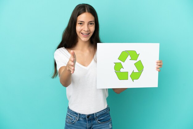 Jovem francesa isolada em um fundo azul segurando um cartaz com o ícone de reciclagem fazendo um acordo