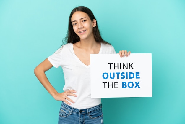 Jovem francesa isolada em um fundo azul segurando um cartaz com o texto Pense fora da caixa com uma expressão feliz