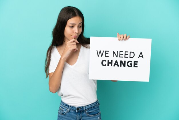 Jovem francesa isolada em um fundo azul segurando um cartaz com o texto Precisamos de uma mudança e pensando