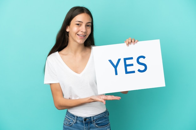 Jovem francesa isolada em um fundo azul segurando um cartaz com o texto SIM com uma expressão feliz